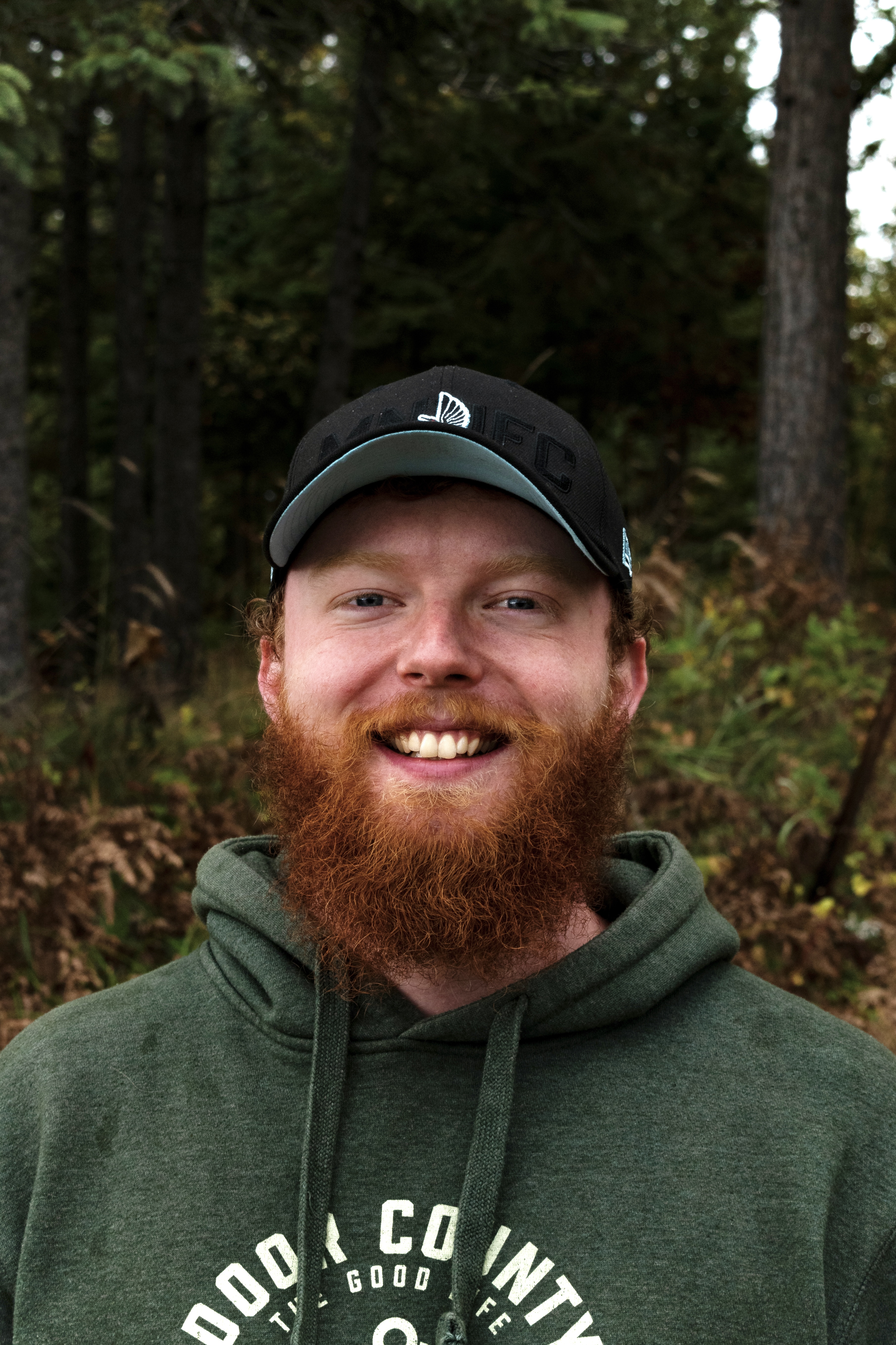 A smiling person with red hair and a beard is wearing a black baseball cap and a green hooded sweatshirt.